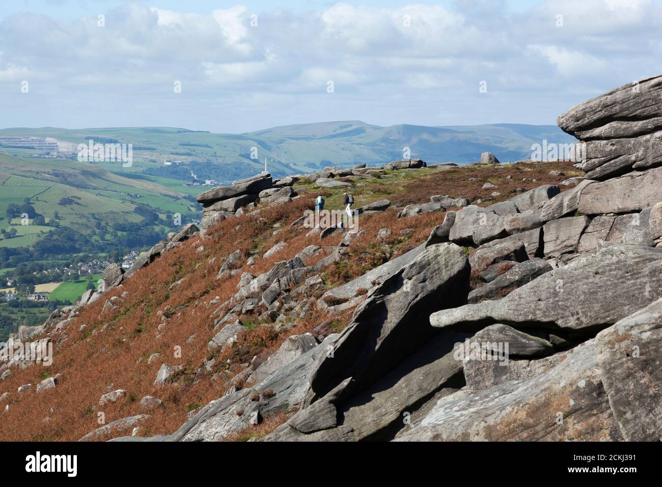Climbers with backpacks walk along the top of a gritstone ridge with sweeping countryside views of the Hope Valley in the Peak District, UK Stock Photo