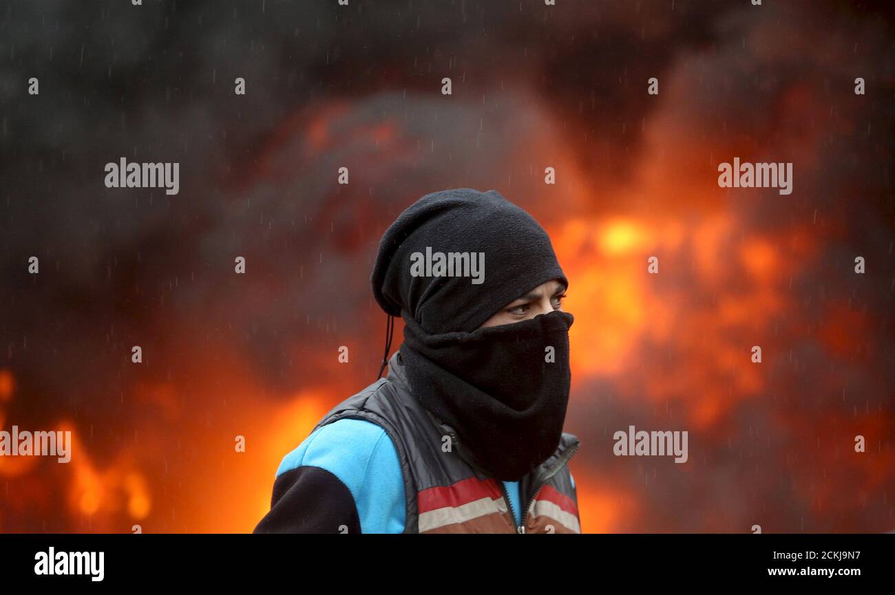 A masked Palestinian protester stands in front of a burning tyre during clashes with Israeli troops in the West Bank town of Qabatya, near Jenin February 6, 2016. On Wednesday, three young Palestinian men from Qabatya wielding guns, knives and pipe-bombs killed a paramilitary Israeli policewoman in Jerusalem and were shot dead. In response, Israeli forces raided the assailants' hometown, arresting five suspected militants and imposing a closure. REUTERS/Mohamad Torokman Stock Photo