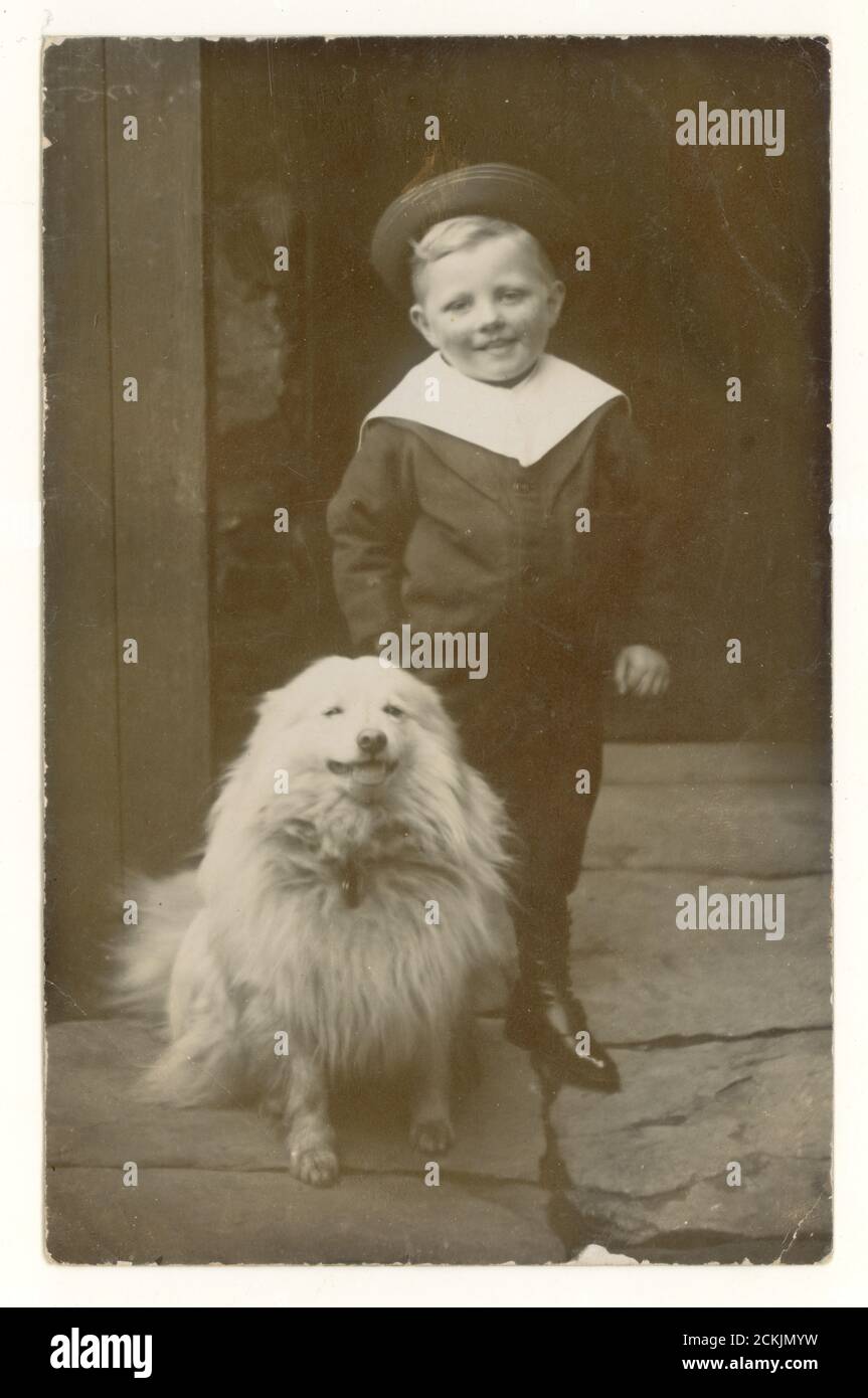 Early 1900's postcard of cute little happy Edwardian boy wearing a sailor suit with his pet dog, possibly the Samoyed breed, standing on flagstones, outside his house, circa 1910, U.K. Stock Photo