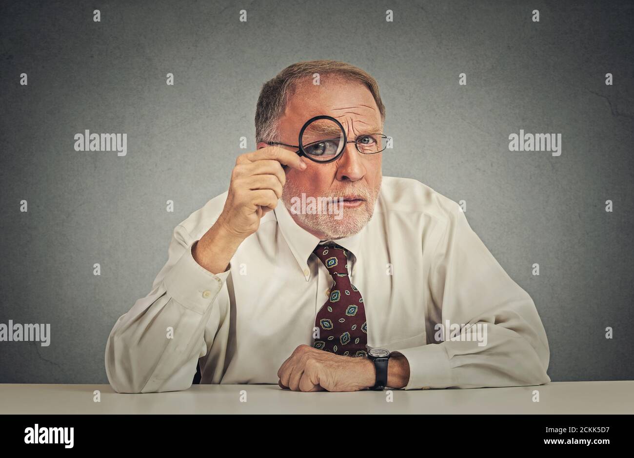 Serious businessman with glasses skeptically looking at you sitting at his office desk isolated on gray wall background. Human face expression, body l Stock Photo