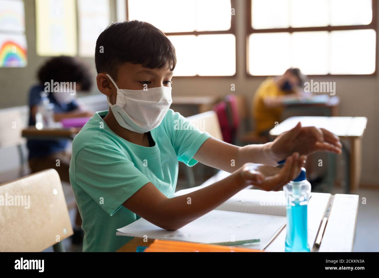 Boy wearing face mask sanitizing his hands while sitting on his desk at school Stock Photo