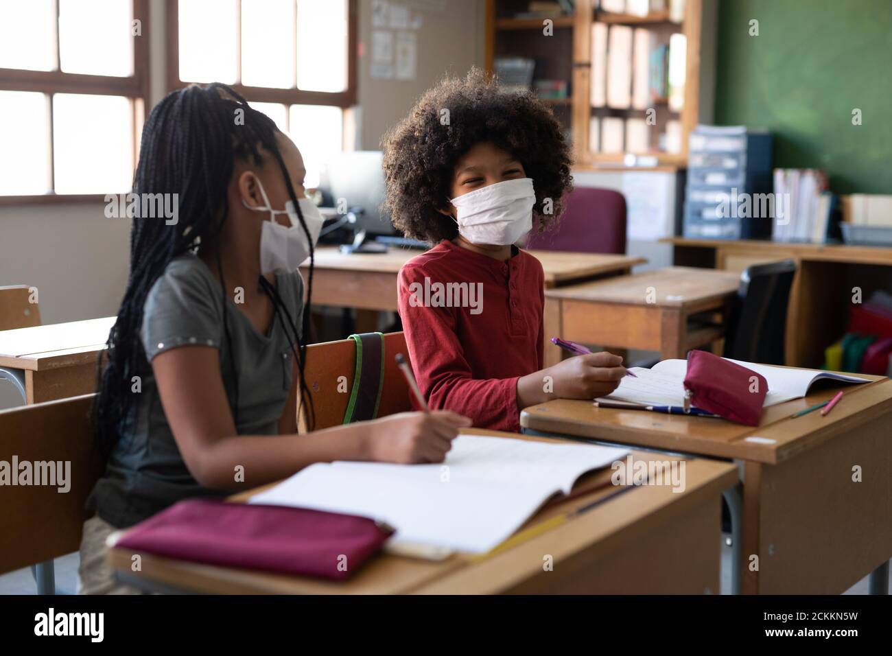 Boy and Girl wearing face masks talking to each other while sitting on their desk at school Stock Photo