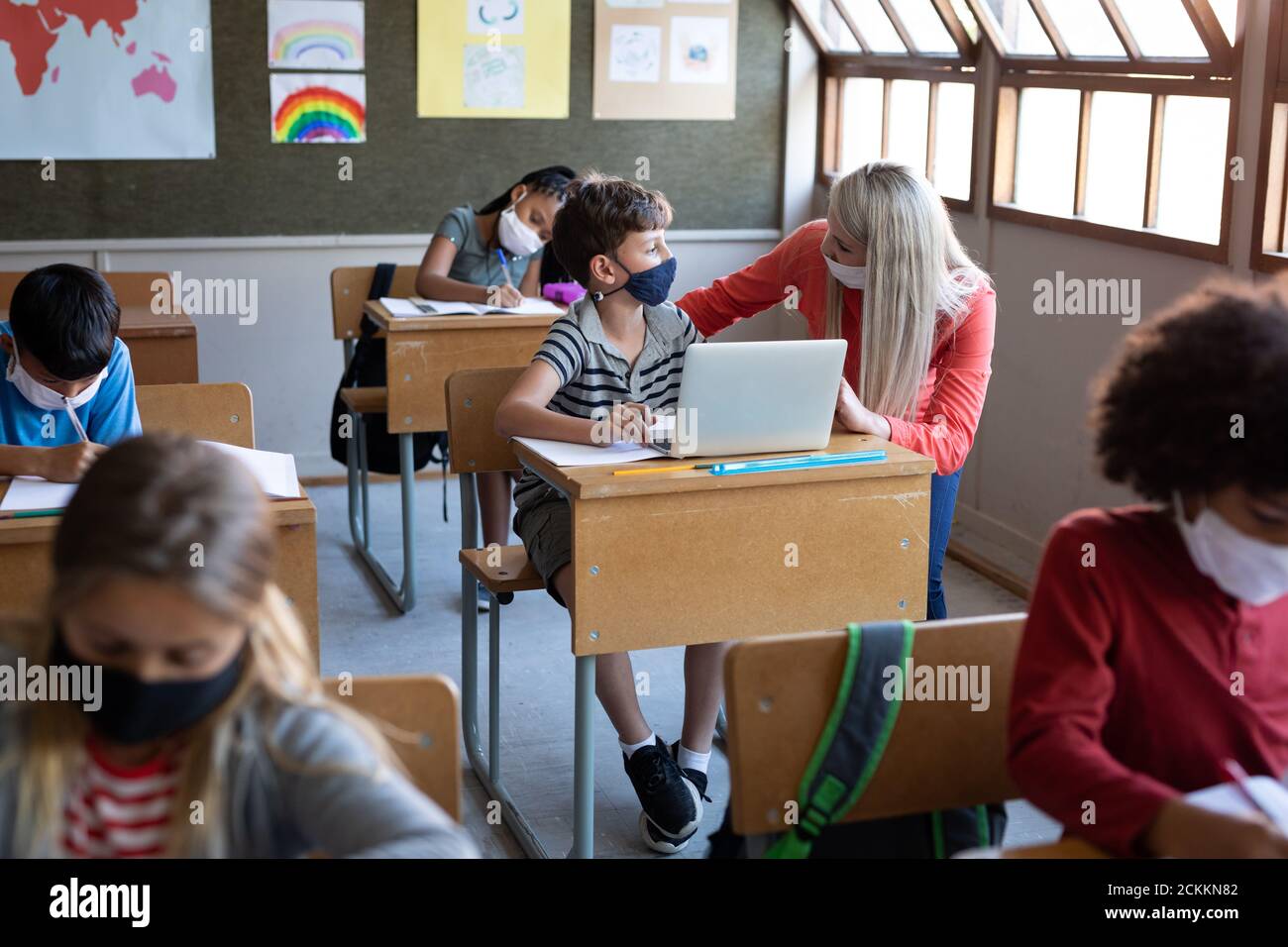 Female teacher and boy wearing face masks using laptop in class at school Stock Photo