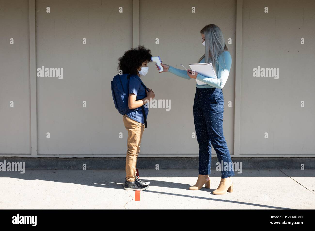 Female teacher measuring temperature of a boy wearing face mask at school Stock Photo