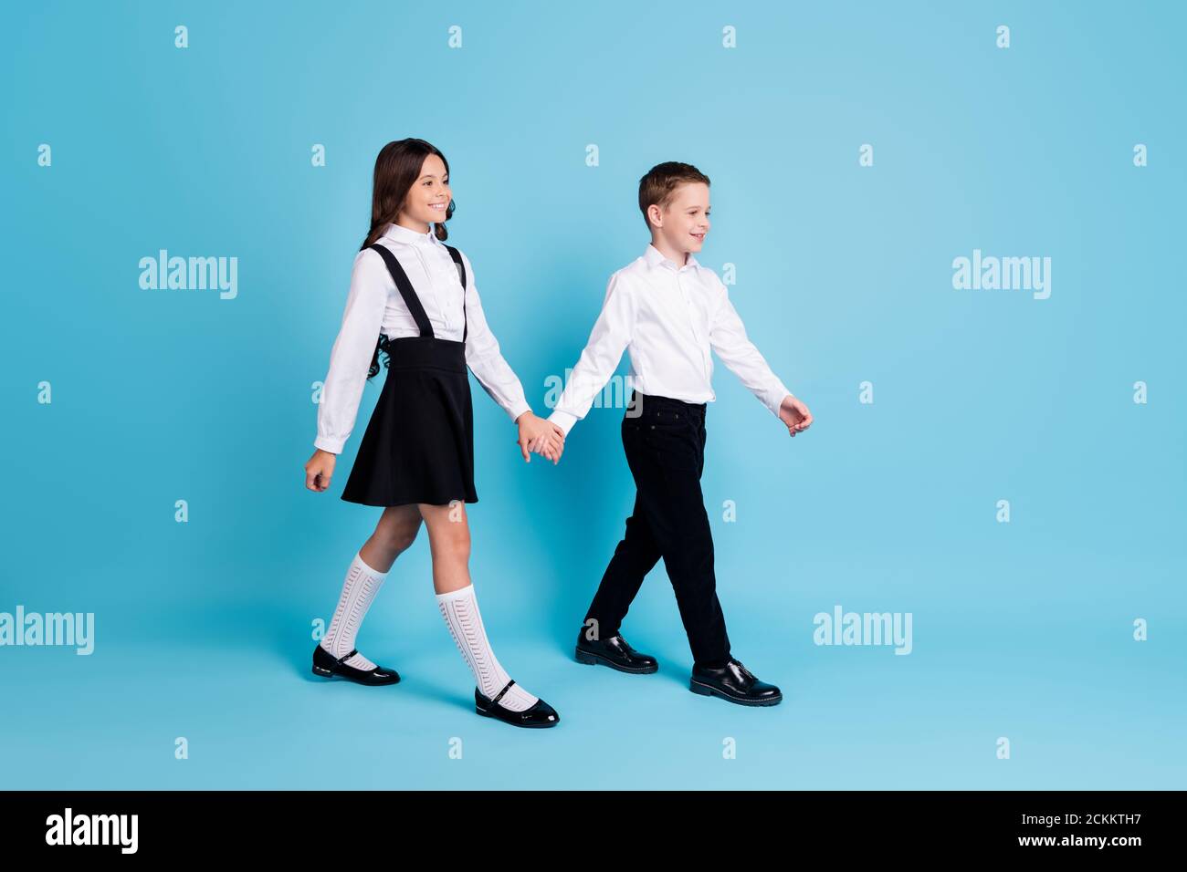 Full length profile photo of two girl boy classmates schoolchildren smiling hold hands morning walk to school wear uniform white shirt black pants Stock Photo