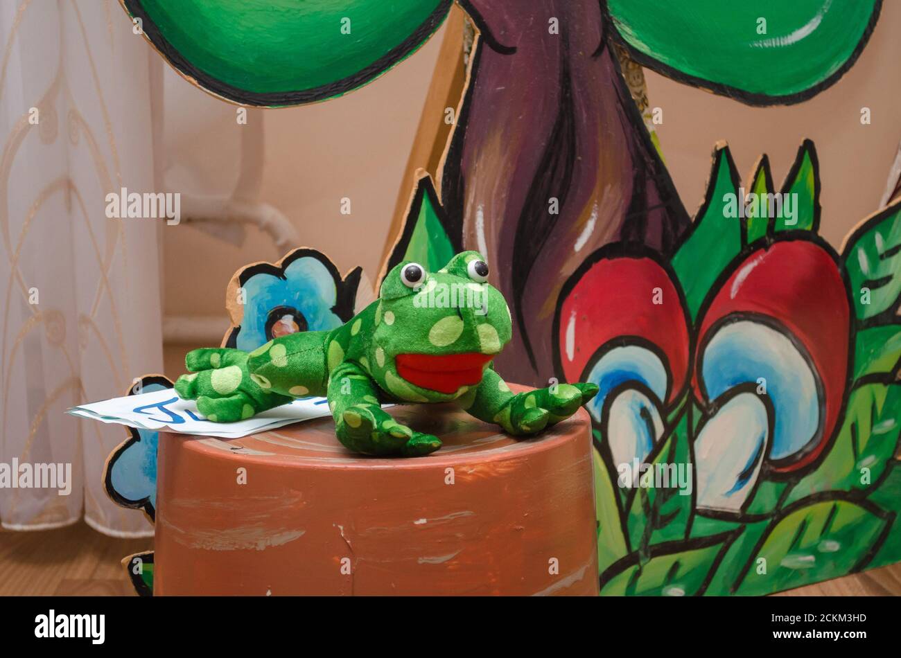 Green toy frog lies on an artificial stump against a background of a drawn tree and mushrooms Stock Photo