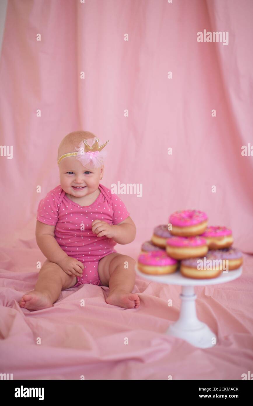 Funny baby girl 1 year old wearing bodysuit and crown headband eating tasty donuts over pink background. Looking at camera. Childhood. Birthday party. Stock Photo