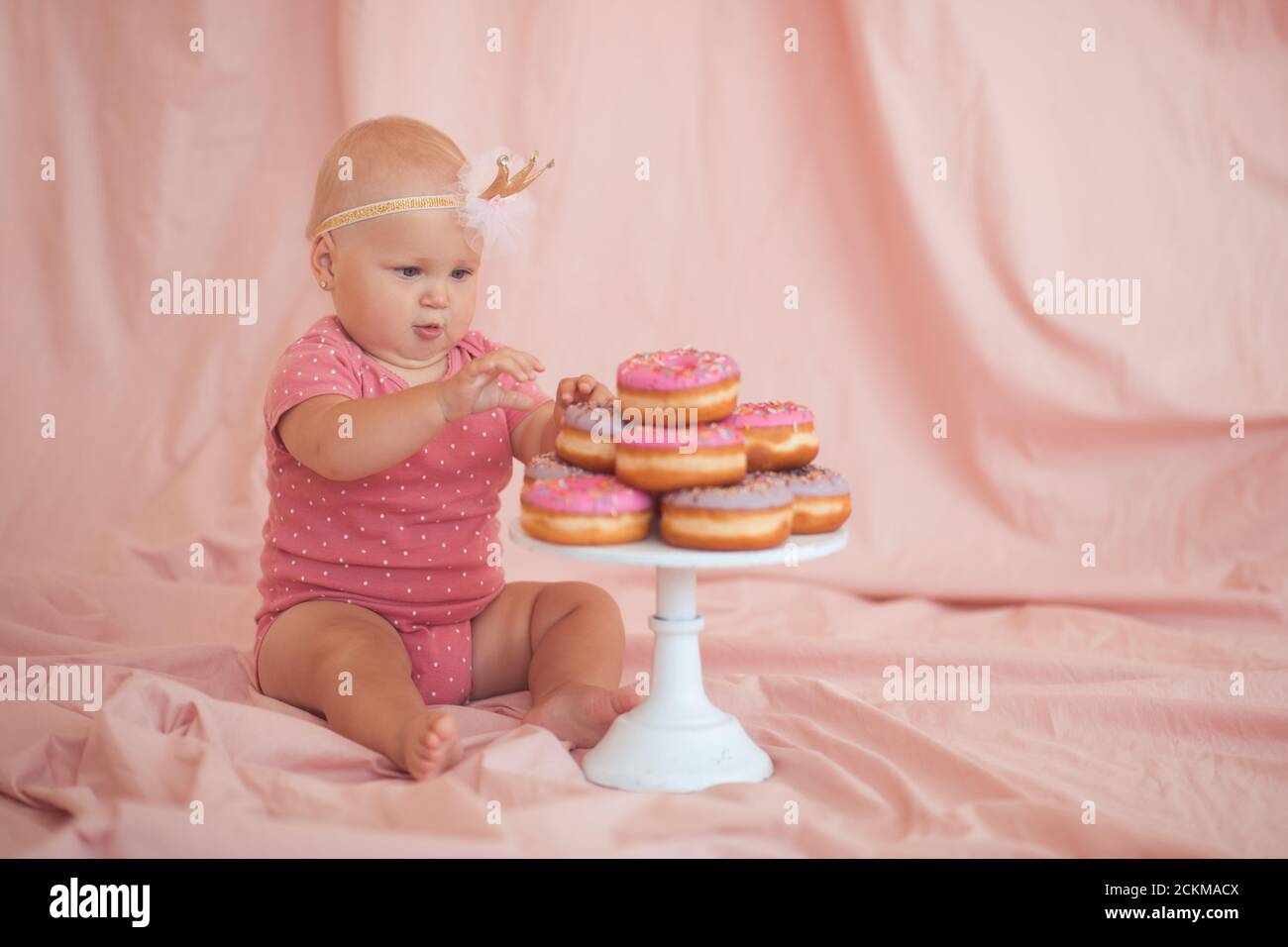 Cute baby girl 1 year old eating tasty glazed donuts sittin on floor over pink textile background closeup. Birthday party. Celebration. Stock Photo