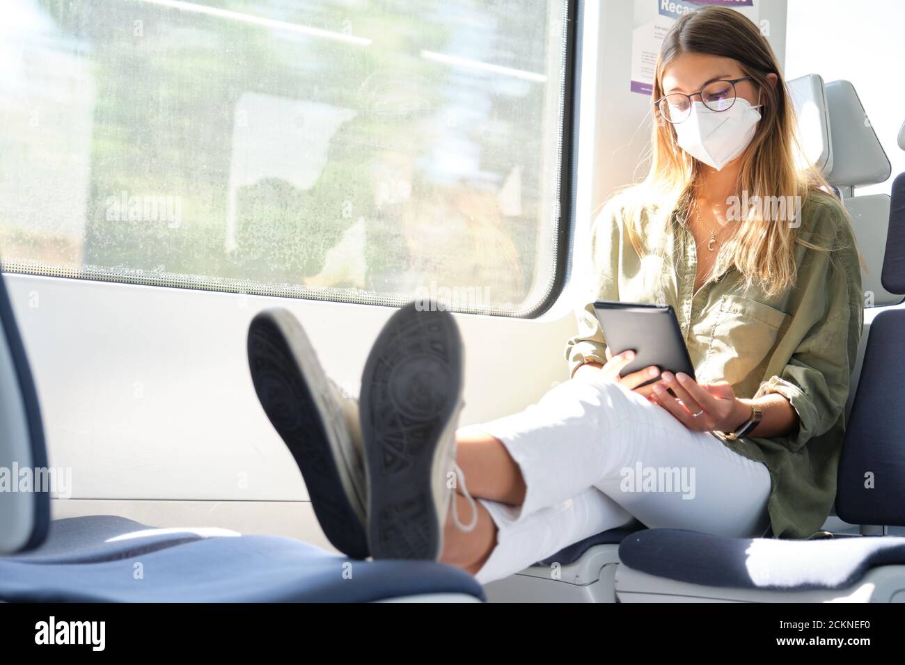 Young woman wearing face mask and reading a e-book in the train with her feet up. New normal travel concept. Stock Photo