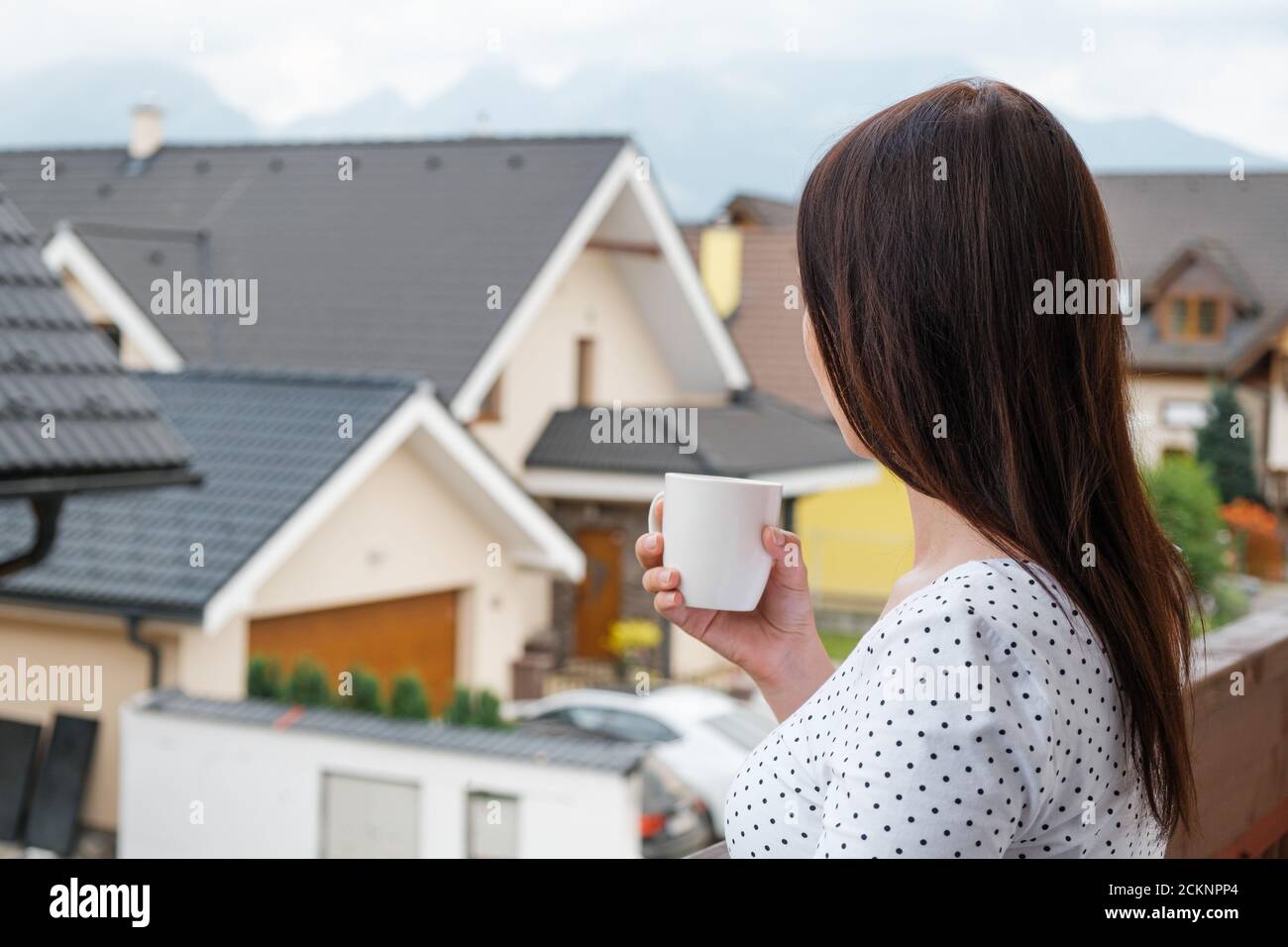 Woman standing with a white cup on the balcony and dreamily watching on the cloudy mountain hill. Morning in the countryside. Modern houses near the Stock Photo