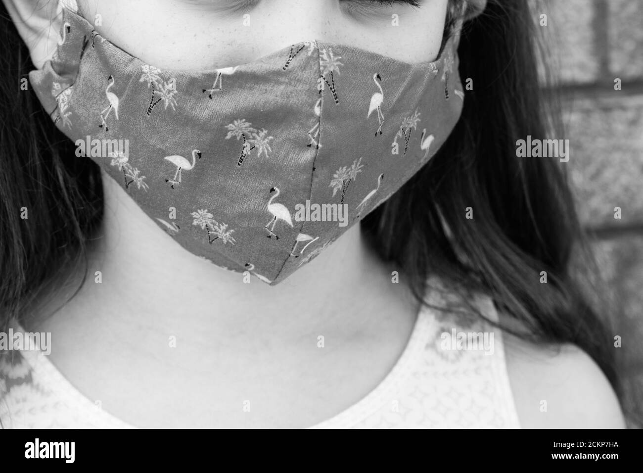 close up of young girl wearing fabric face mask outside with brick wall background in sunshine. Face covering used for shielding Stock Photo