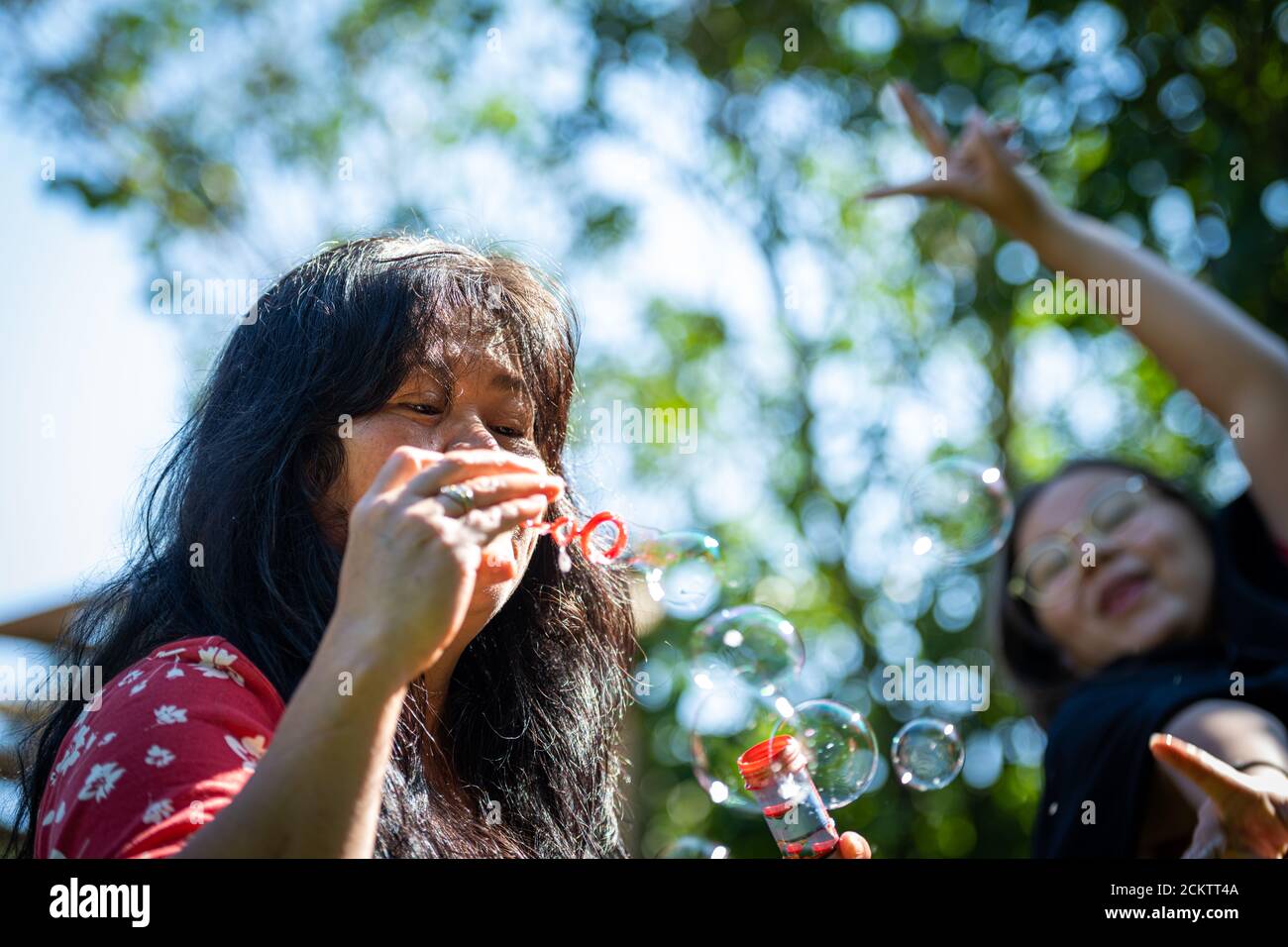 Closeup picture of a middle-aged, in her 50s, Asian woman with soap bubbles. A preteen girl and green leaves in the background Stock Photo