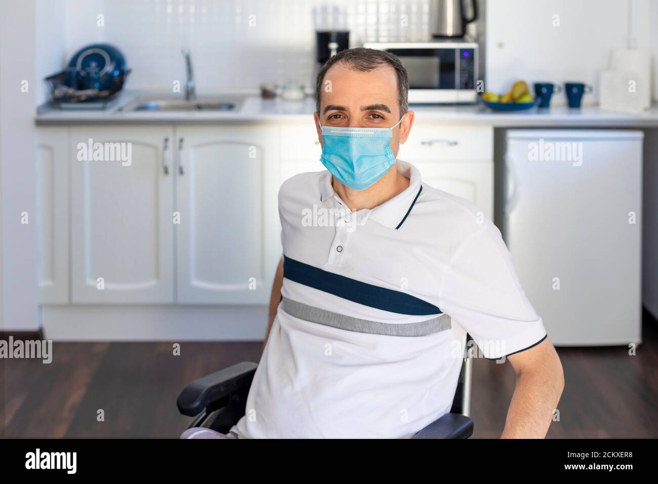 Smiling Young Handicapped Man Sitting On Wheelchair In Kitchen.  Young man wearing face mask sitting infront of kitchen. Focus on his face. Stock Photo