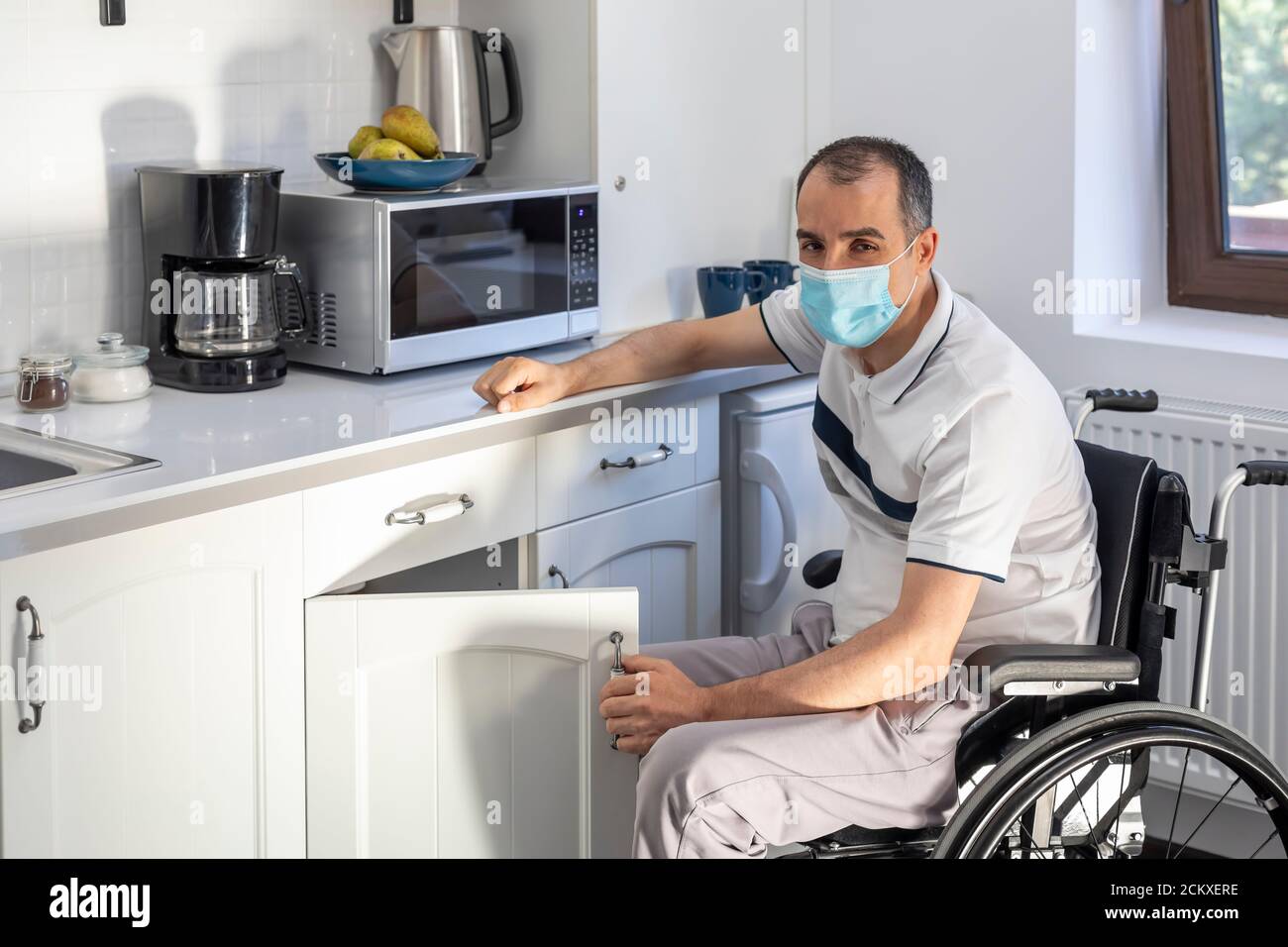 Smiling Young Handicapped Man Sitting On Wheelchair In Kitchen.  Young man wearing face mask sitting in front of kitchen. Focus on his face. Stock Photo