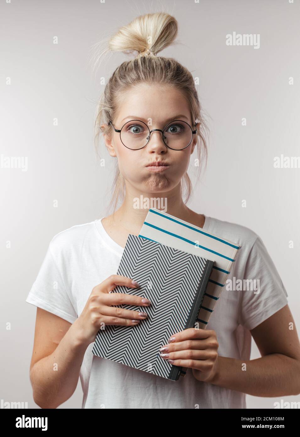 Tired young blonde female student puffing her cheeks holding several books in hands and looking at camera isolated over gray wall background. Stock Photo