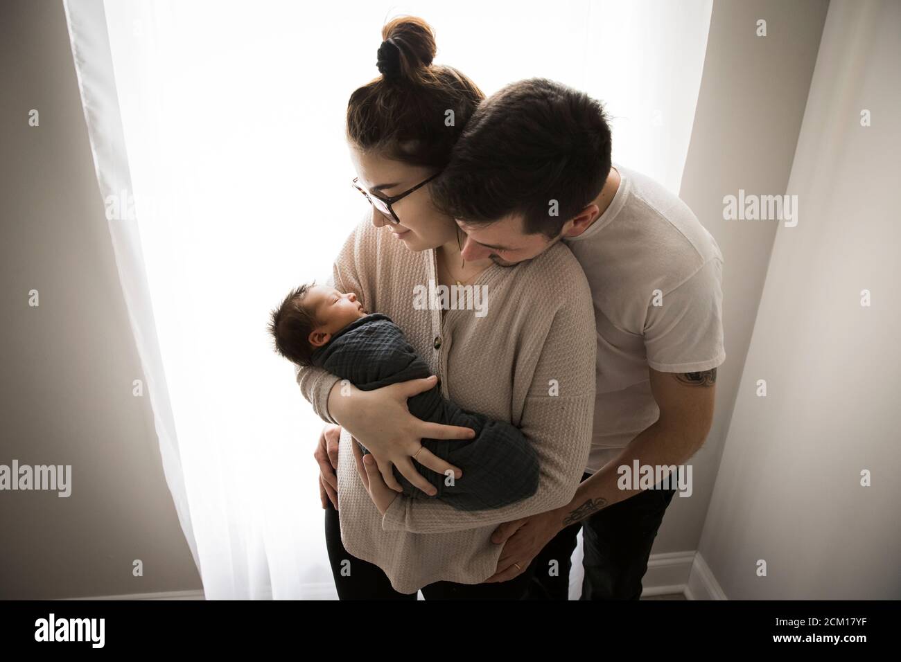 Gen Z Parents Snuggle by Window and Admire Their Newborn Baby Stock Photo