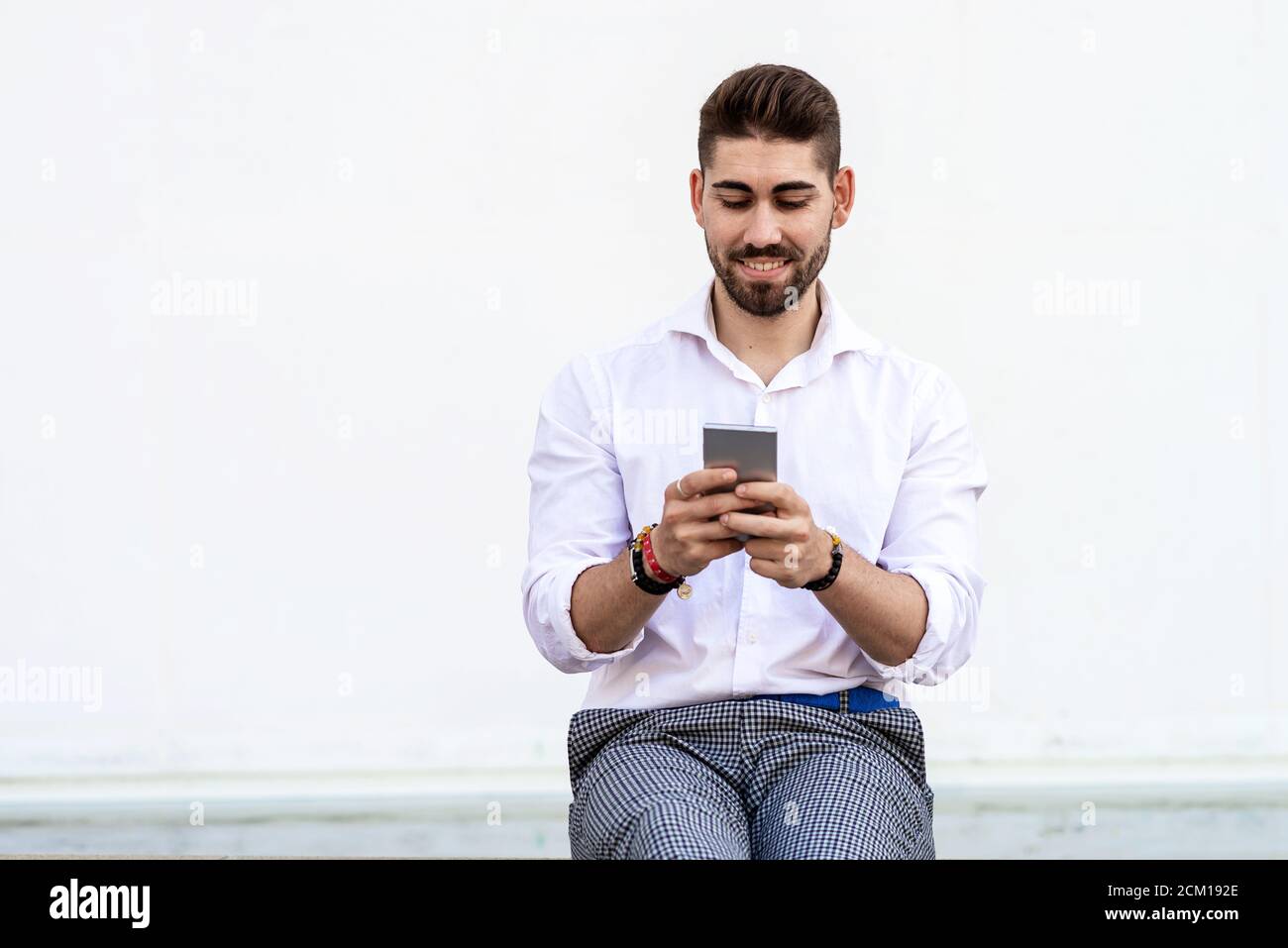 Young bearded man sitting against a white wall using the mobile phone Stock Photo