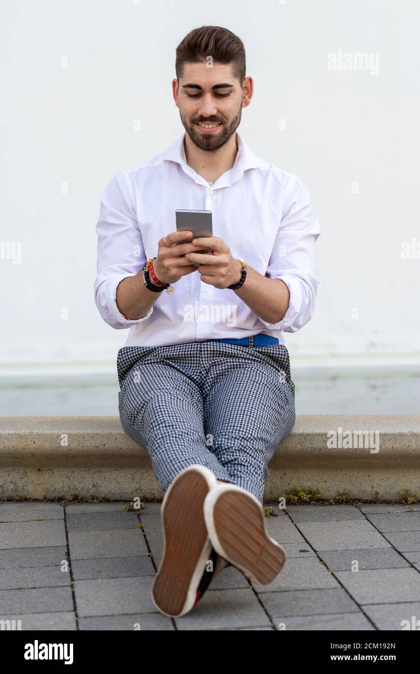 Young bearded man sitting against a white wall using the mobile phone Stock Photo