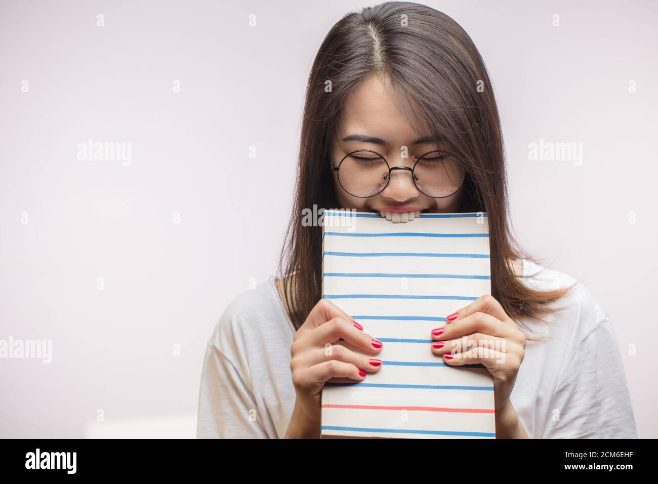 Excited playful Asian girl with dark hair, bites a book , looks nervously at camera, wears spectacles, uses literature for learning material, stands i Stock Photo