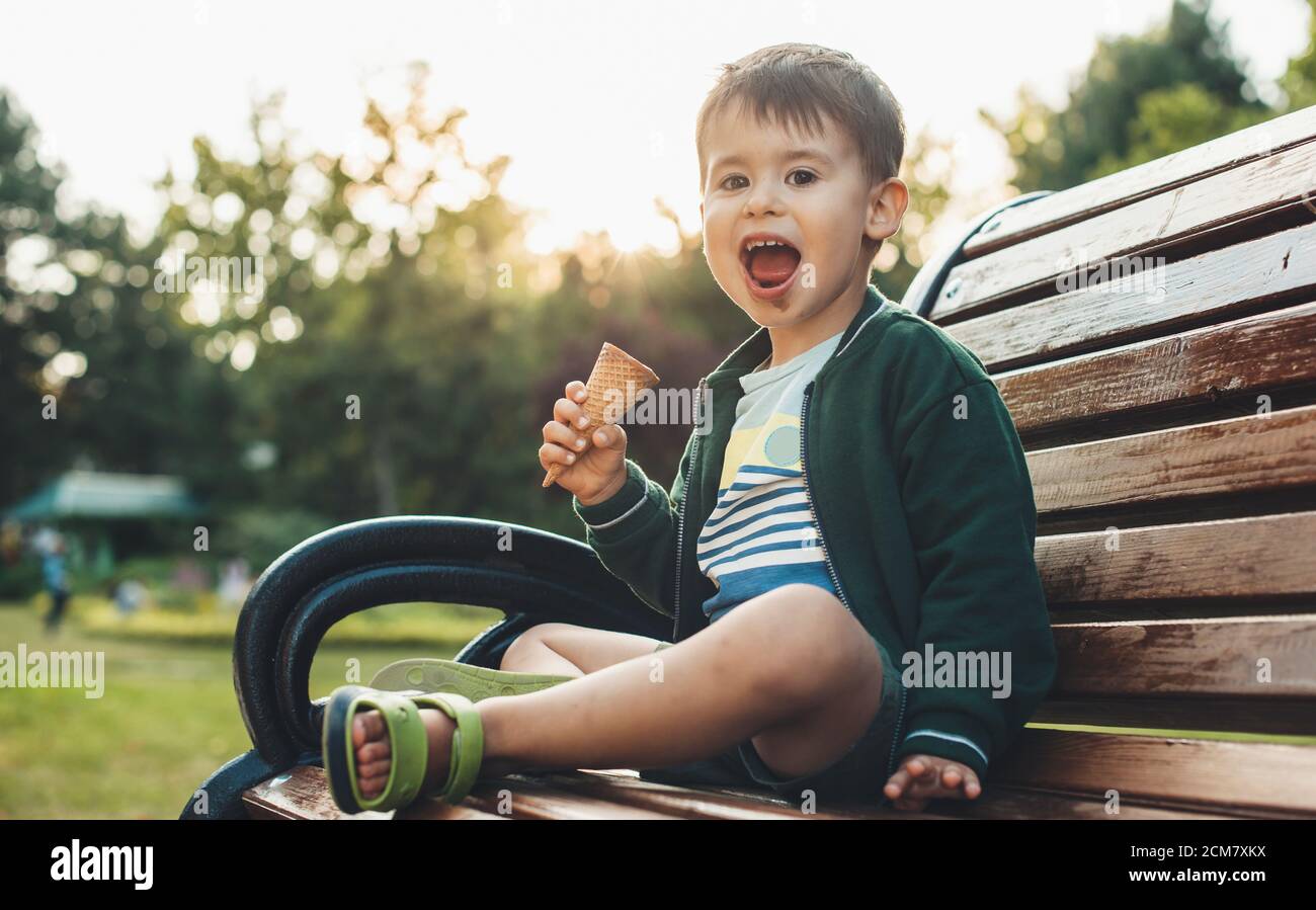 Caucasian boy sitting in the bench in park is eating an ice-cream and smile at camera with dirty mouth Stock Photo