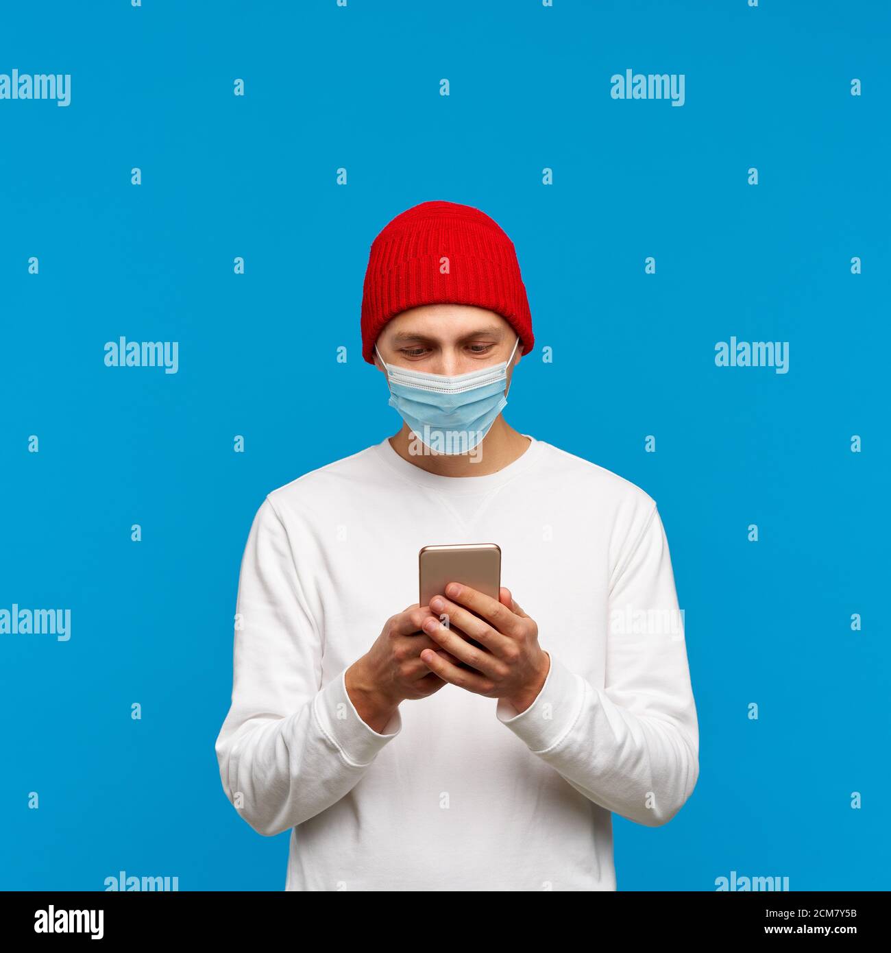 Portrait of man with medical protective face mask, texting on mobile phone. Male write message isolated on bright blue colored background. White jumper and red hat. Stock Photo