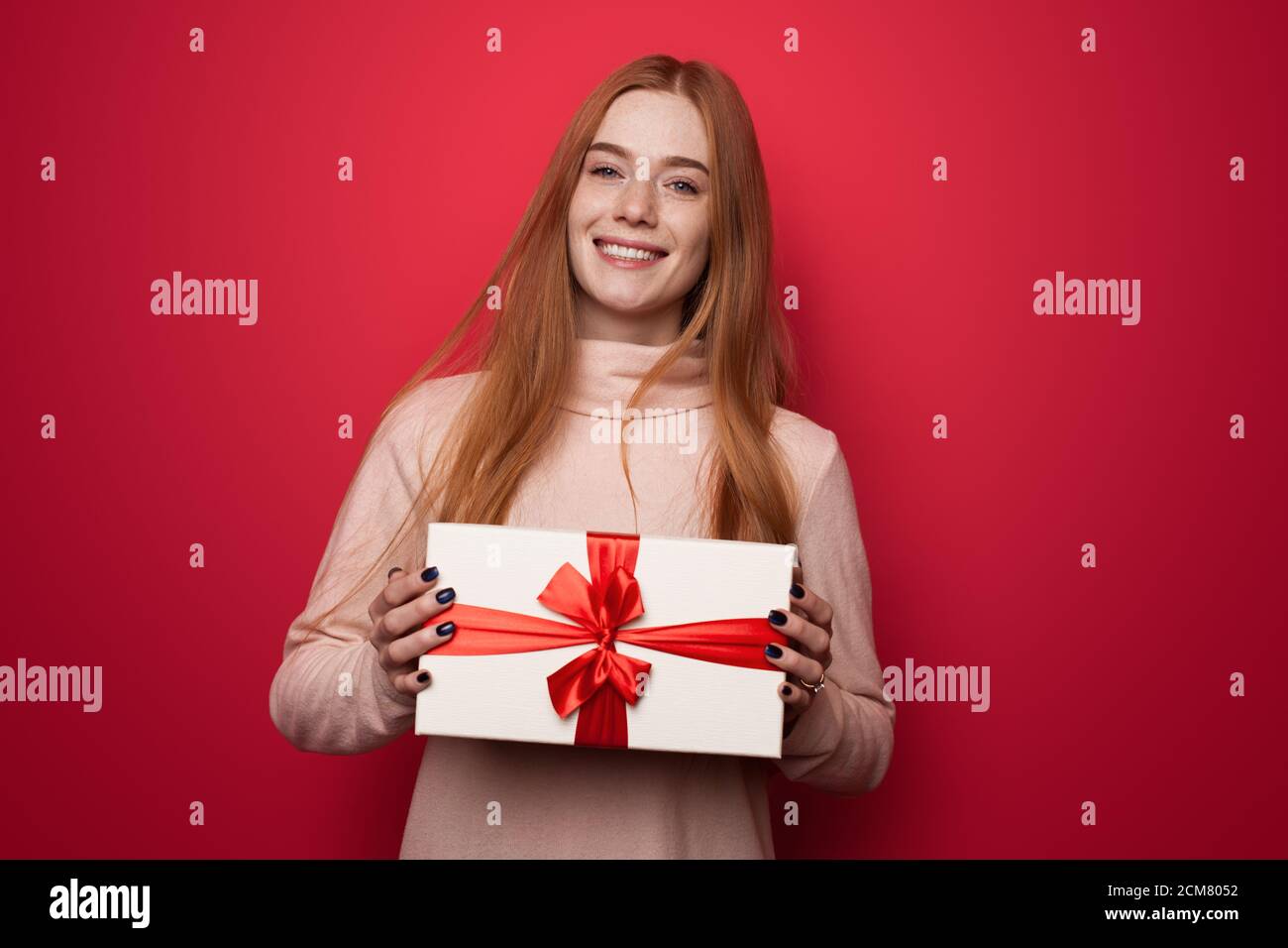 Beautiful caucasian woman with red hair and freckles smiling at camera and holding a present in hands posing on a red studio wall Stock Photo