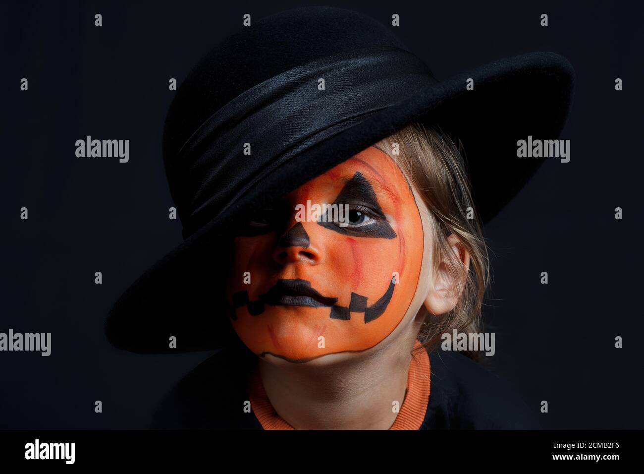 sad child with a pumpkin pattern on his face in the black hat on a black background, Halloween and looks like a Joker Stock Photo