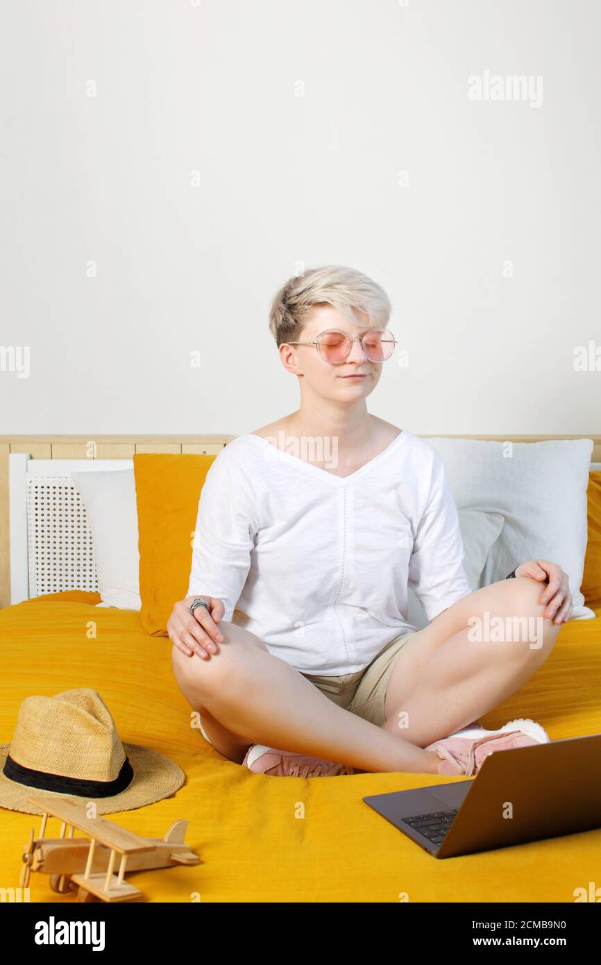 Blonde pretty hipster young woman meditating online sitting on a bed in front of laptop during daybreak, dreaming about travels and adventures Stock Photo