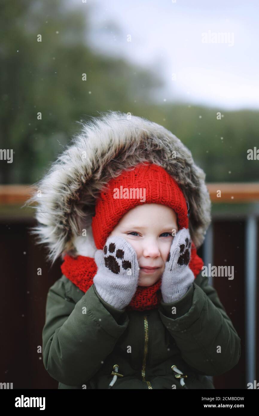 Portrait of cute sweet little girl in warm outerwear clothing and hood outdoors in cold snowy weather in wintertime, funny mittens with paws Stock Photo