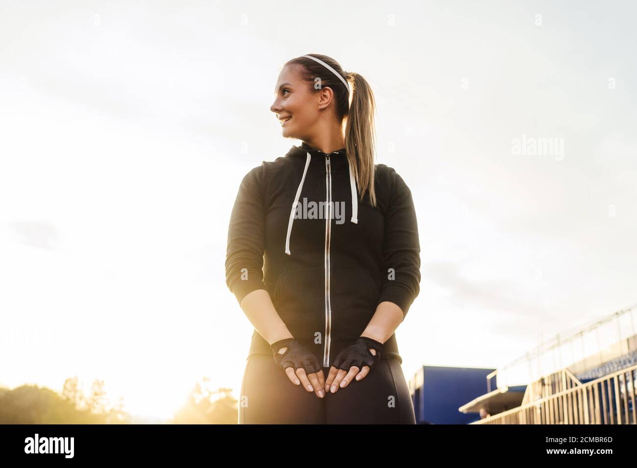 Fitness smiling woman portrait lifestyle in black tracksuit outdoor, white sunny sky background. Sunset light. Stock Photo