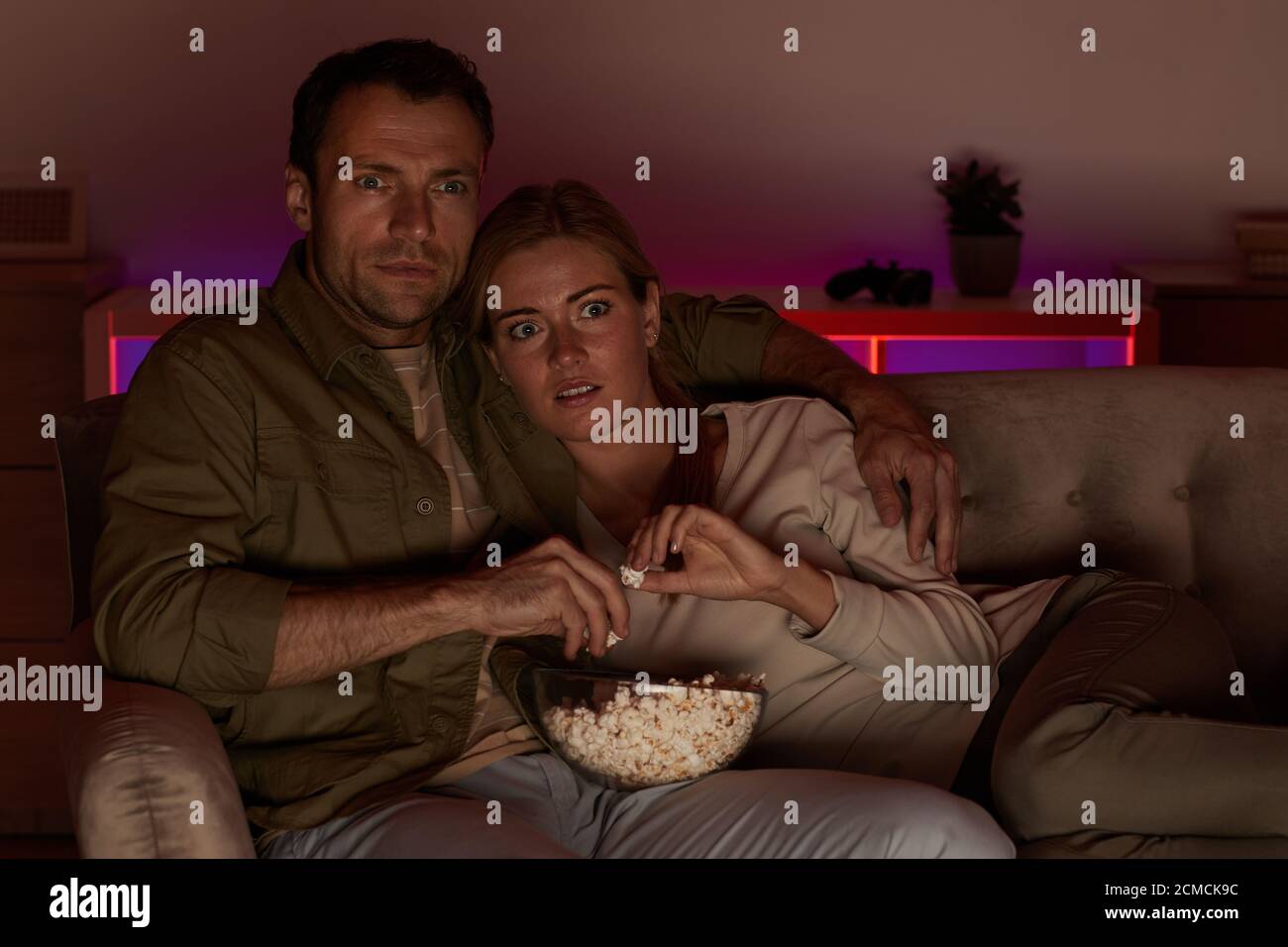 Young couple looking very frightened they lying on sofa with popcorn and watching scary movie Stock Photo
