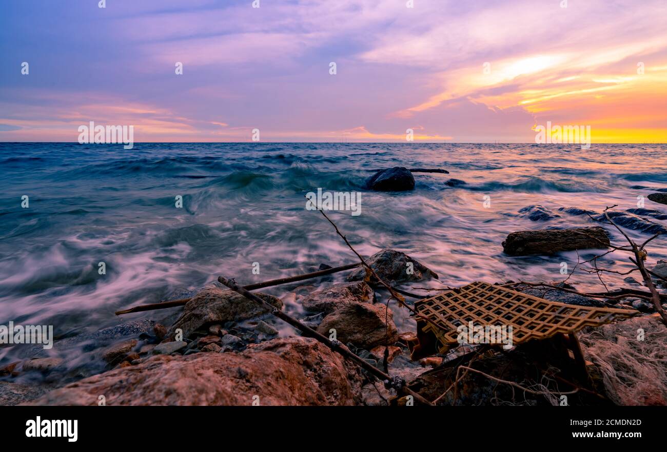 Sea water with golden and purple sunset sky. Plastic waste on rock beach. Tropical beach and ocean pollution. Ocean water. Seashore on summer. Stock Photo