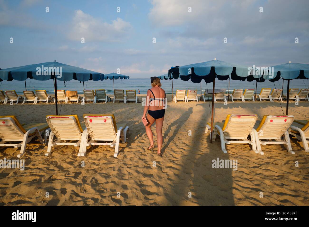 In early morning light, a lone Western female tourist walks between the umbrellas and deck chairs set up on Karon Beach, Phuket, Thailand Stock Photo