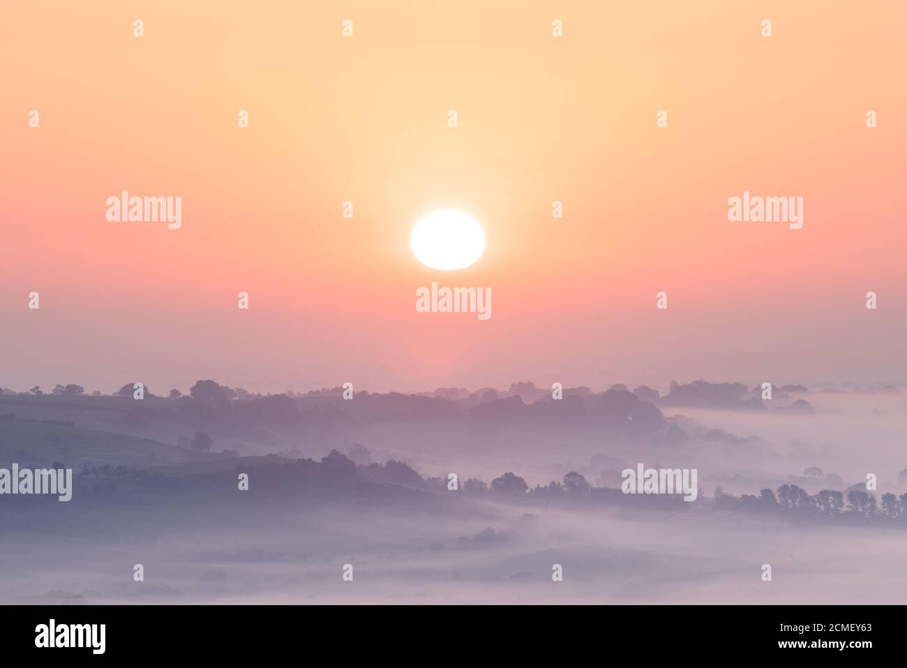 The rising sun casts pastel tones across the fog shrouded landscape of Lower Wharfedale, with tree lined hills peeking through the mist. Stock Photo