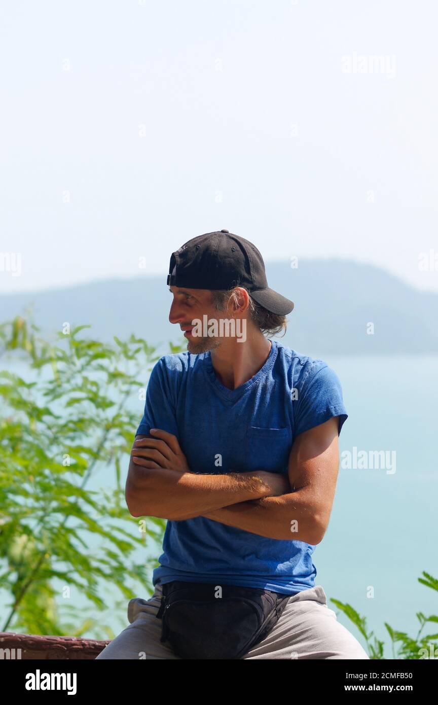 Happy Young Bearded Man sitting on the stone railings in city park wearing in blank blue t-shirt and black Cap Stock Photo