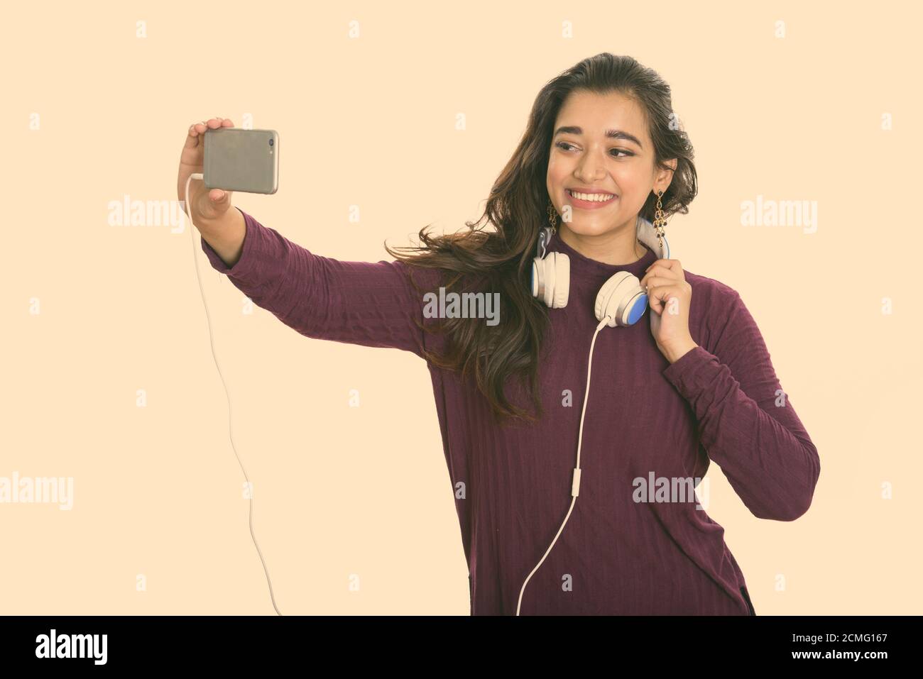 Young happy Indian woman smiling while wearing headphones around neck and taking selfie picture with mobile phone Stock Photo