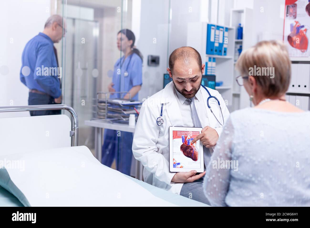 Doctor explaining coronary artery disease to senior woman patient pointint at tablet pc in hospital examination room. Medical services medicine healthcare well-being support, physician working Stock Photo