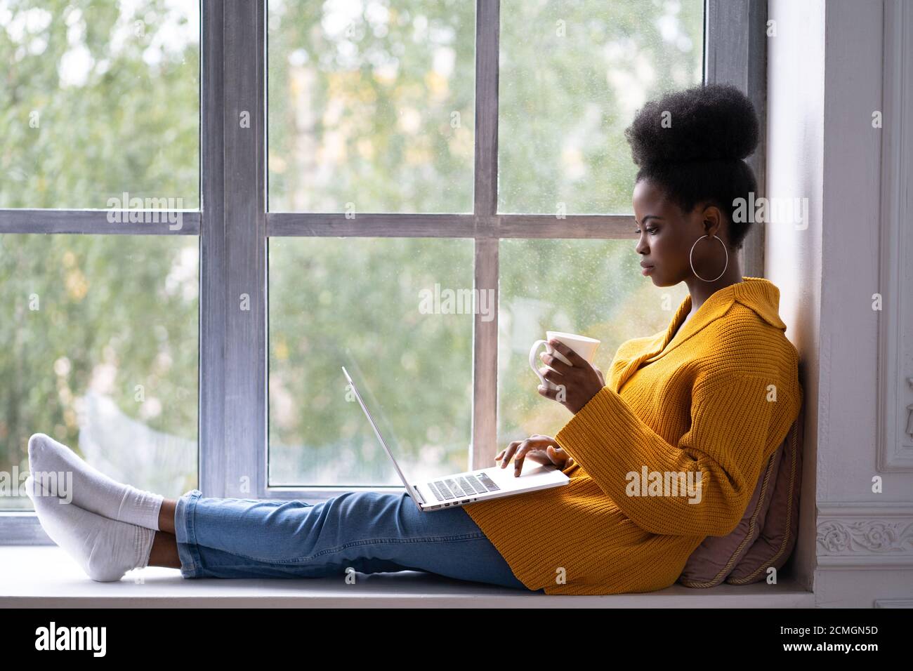 Focused African American woman student with afro hairstyle wear yellow cardigan, sitting on windowsill, working doing remote job on laptop, preparing Stock Photo