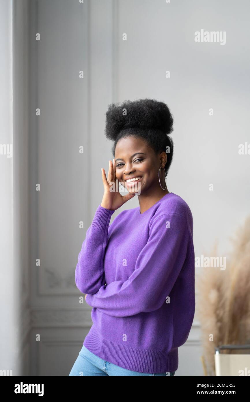 Portrait of smiling african American millennial young woman in trendy jewelry big earrings wear purple sweater standing and posing at home. Cheerful g Stock Photo