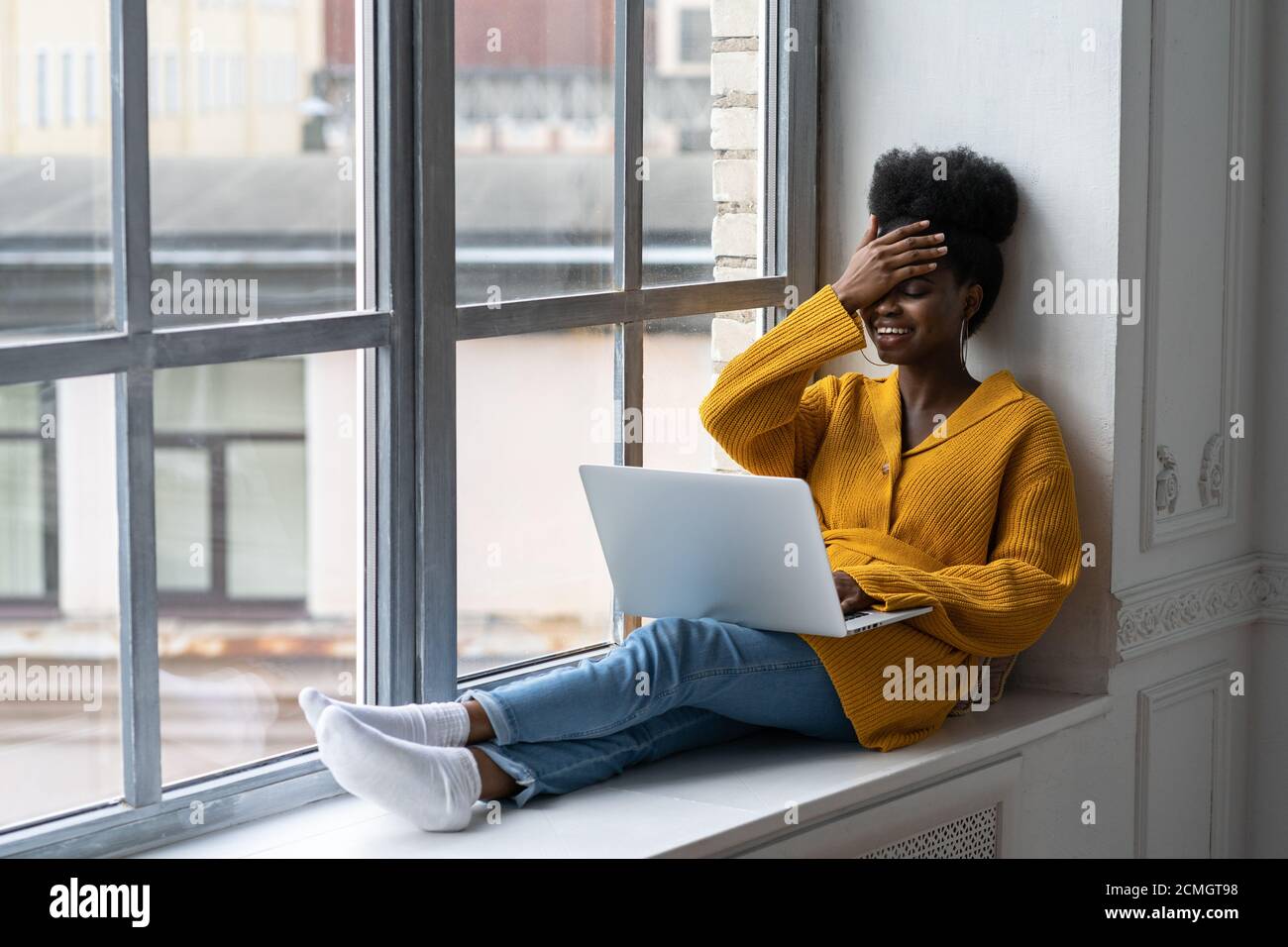 Happy African American woman freelancer with afro hairstyle wear yellow cardigan sitting on windowsill, working on laptop, talking in video chat, laug Stock Photo