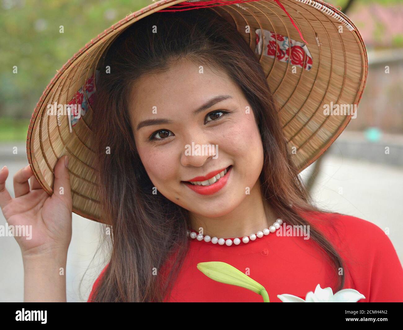 Young Vietnamese woman in red wears a traditional Asian conical straw hat, holds the brim of the hat with her right hand and poses for the camera. Stock Photo