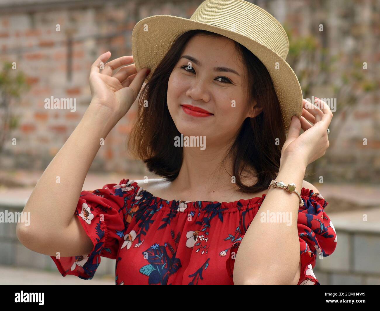 Beautiful young Vietnamese woman wears an Amish-style straw hat, holds the brim of the hat with both hands, and poses for the camera. Stock Photo