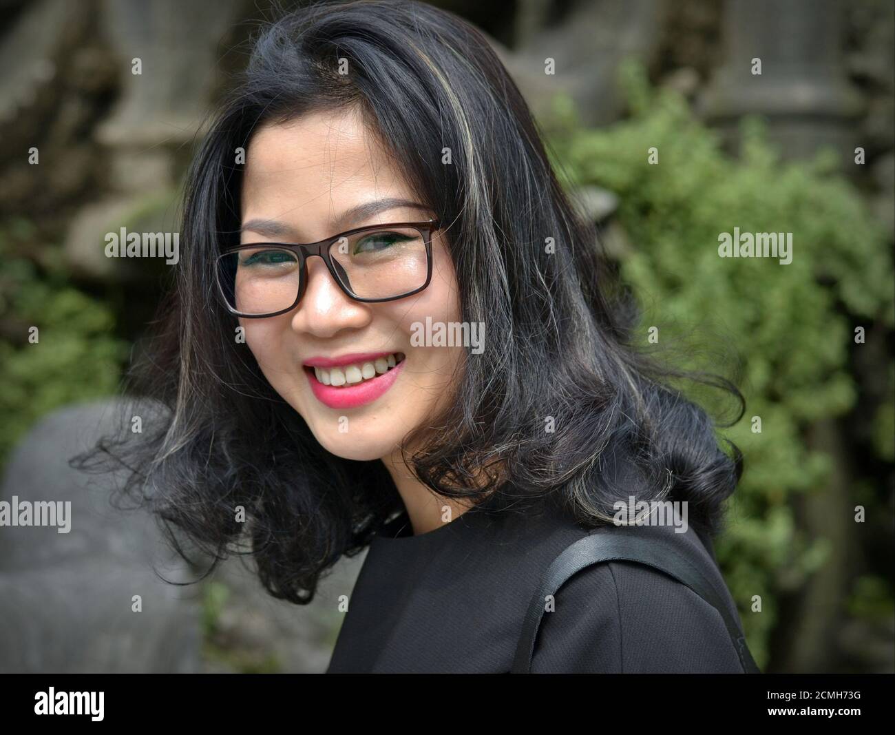 Young attractive cheerful Vietnamese woman with large eyeglasses wears a black top and smiles for the camera. Stock Photo