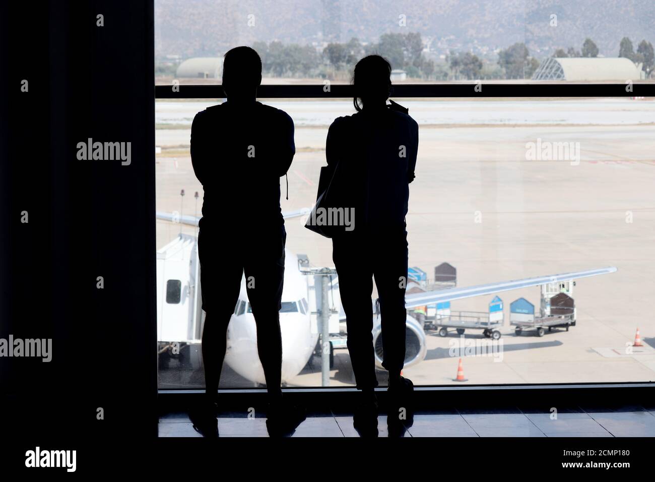 Couple of passengers in the airport, silhouettes of a man and woman in masks looking at the plane on the tarmac through the glass Stock Photo