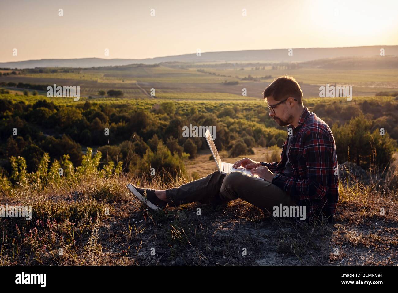 man with laptop sitting on the edge of a mountain with stunning views of the valley. Cellular network broaband coverage concept. 5G. Stock Photo