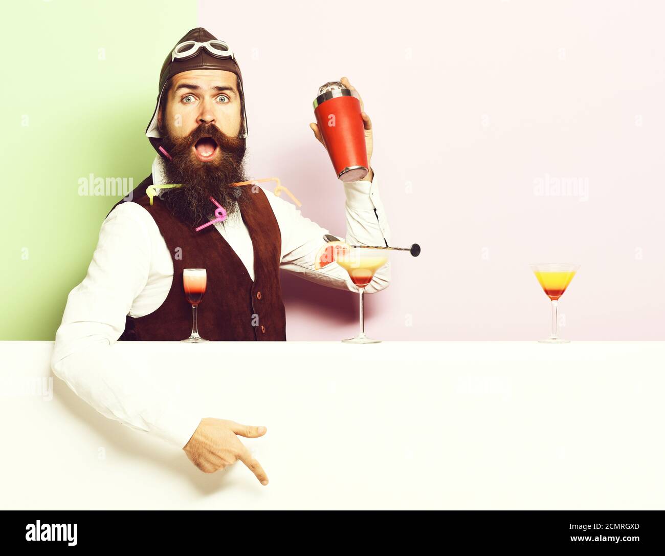 handsome bearded pilot with long beard and mustache on surprised face with shaker alcoholic cocktails in vintage suede leather waistcoat with hand and glasses, on purple green studio background Stock Photo
