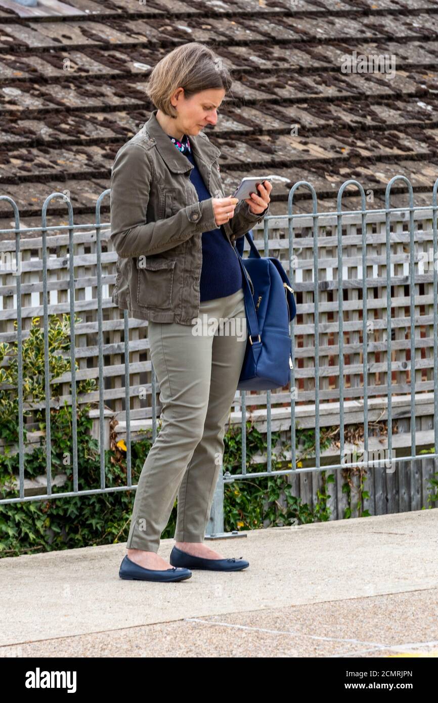 a middle-aged woman waiting for a train on a railway platform using a mobile device or phone to check the timetable. Stock Photo