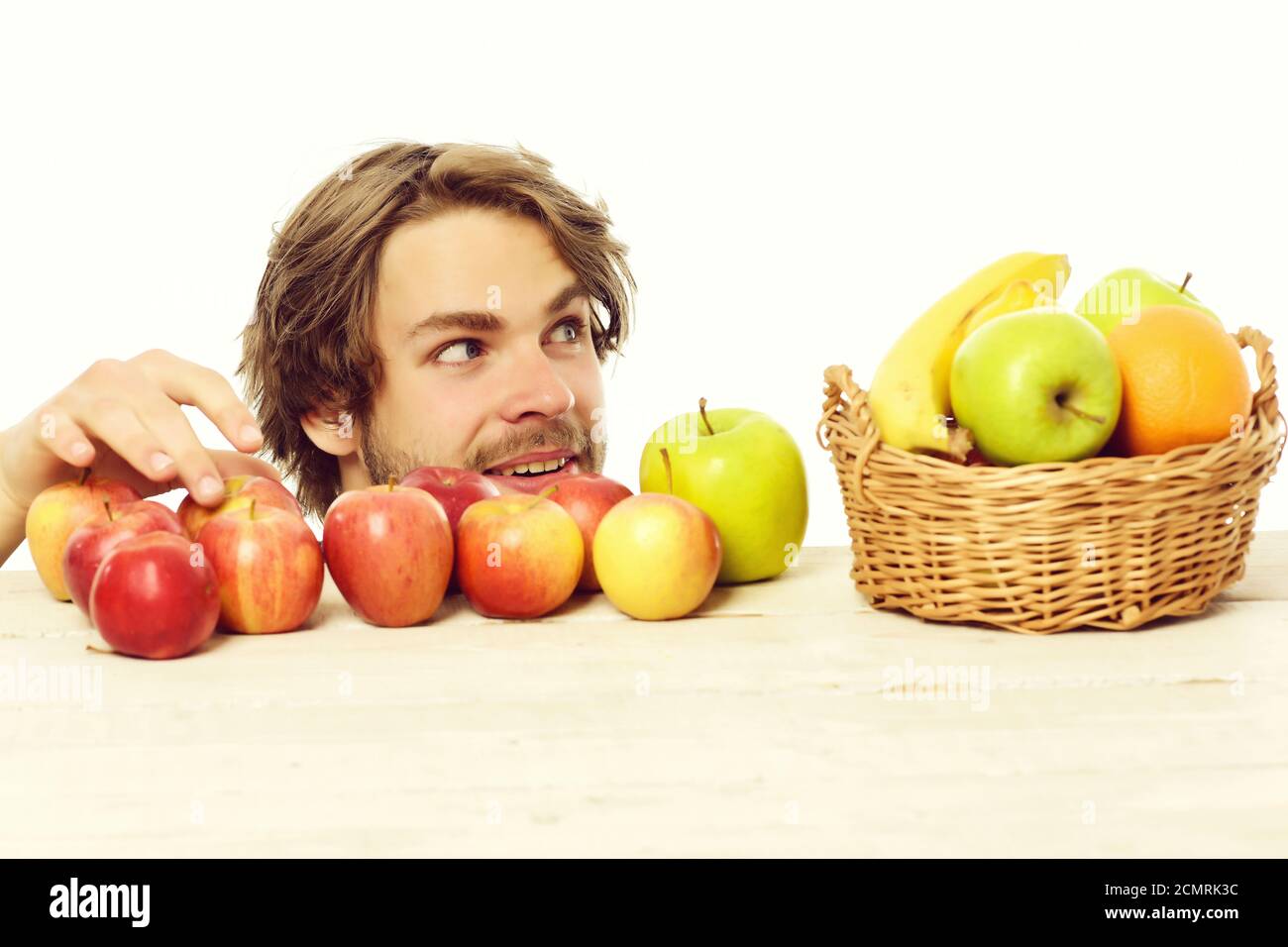 Man stealing apple from wooden table near basket of tropical fruit and green apples on white background Stock Photo