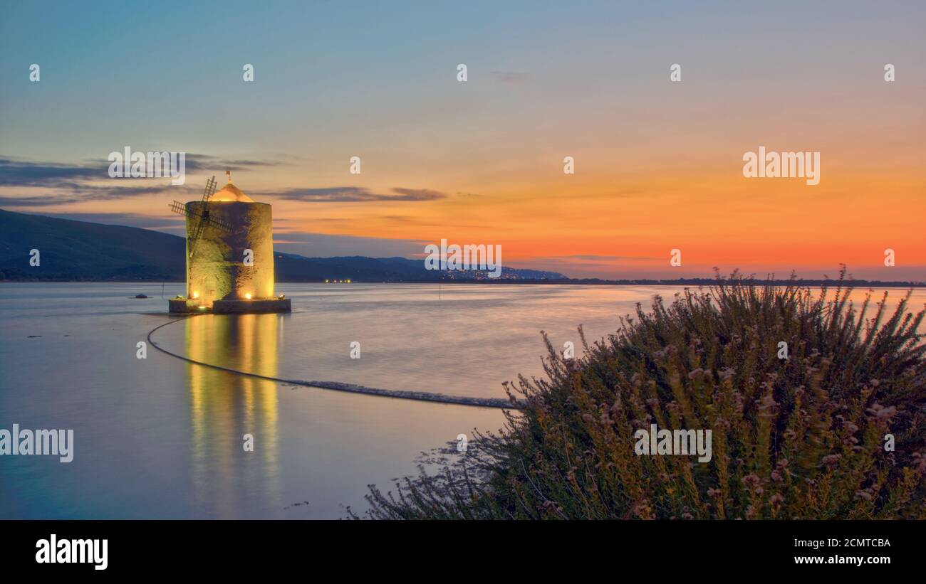 Orbetello lagoon with its iconic mill and a heather bush illuminated by the warm light of the sunset Stock Photo
