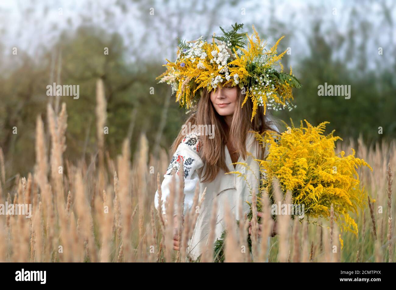 Beautiful girl in a wreath and a ancient embroidered clothes gathers wildflowers in a meadow. Ukrainian culture, ethno style Stock Photo
